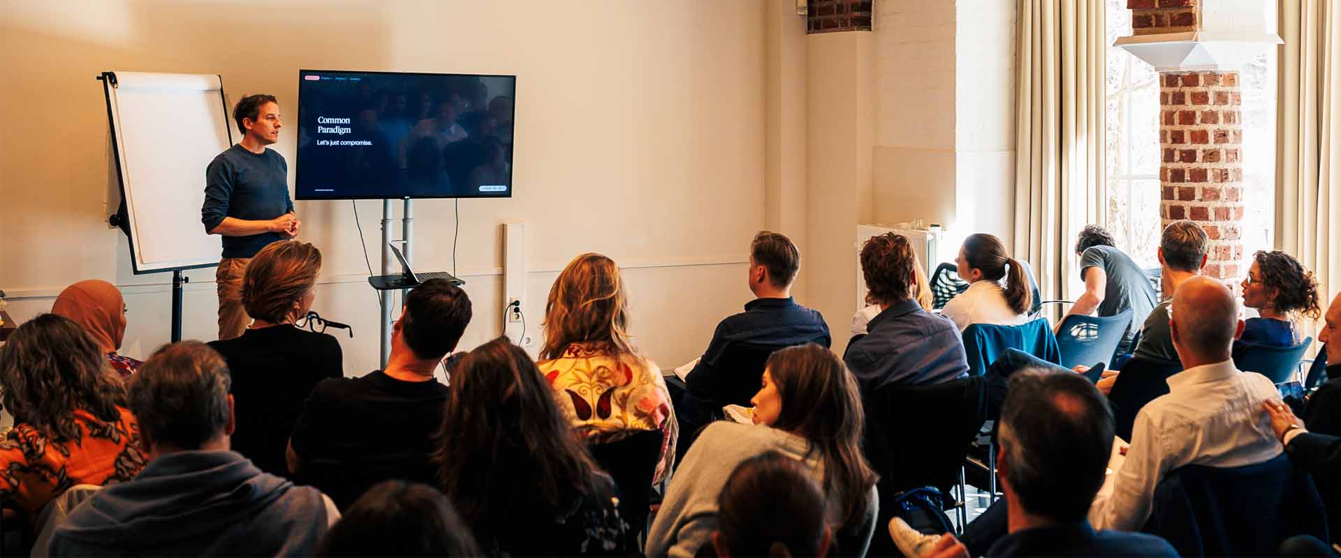 Trainer naast een whiteboard en scherm die voor een groep zittende deelnemers een training geeft